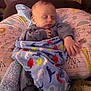 baby, sleeping, blanket, dinosaur_pattern, pillow, animal_illustrations, cozy, indoors, child, infant, soft_lighting, peaceful, resting, cute, clothing, hand, foot, nursery, comfort, portrait