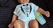 Silas is registered to the contest to win money with this photo: baby, infant, child, smiling, portrait, sitting, onesie, bottle, baby_bottle, black_background, couch, velvet, indoor, closeup, face, hands, feet, happy, casual, photography