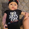 baby, black_beanie, black_tshirt, casual_clothing, child, closeup, curious, cute, expression, face, foot, hand, headwear, indoor, infant, lying_down, person, portrait, skin, text_on_clothing