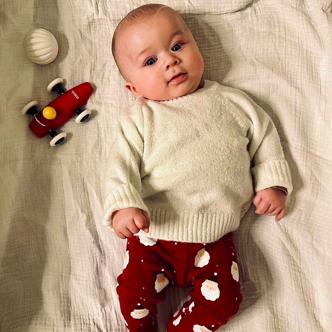 Ezio a rejoint le concours — aidez-le/la à gagner de superbes lots ! baby, blanket, child, cute, expression, face, feet, fingers, holiday_theme, indoors, infant, looking_at_camera, lying_down, playtime, red_pants, santa_print, soft_texture, sweater, toy_car, white_ball