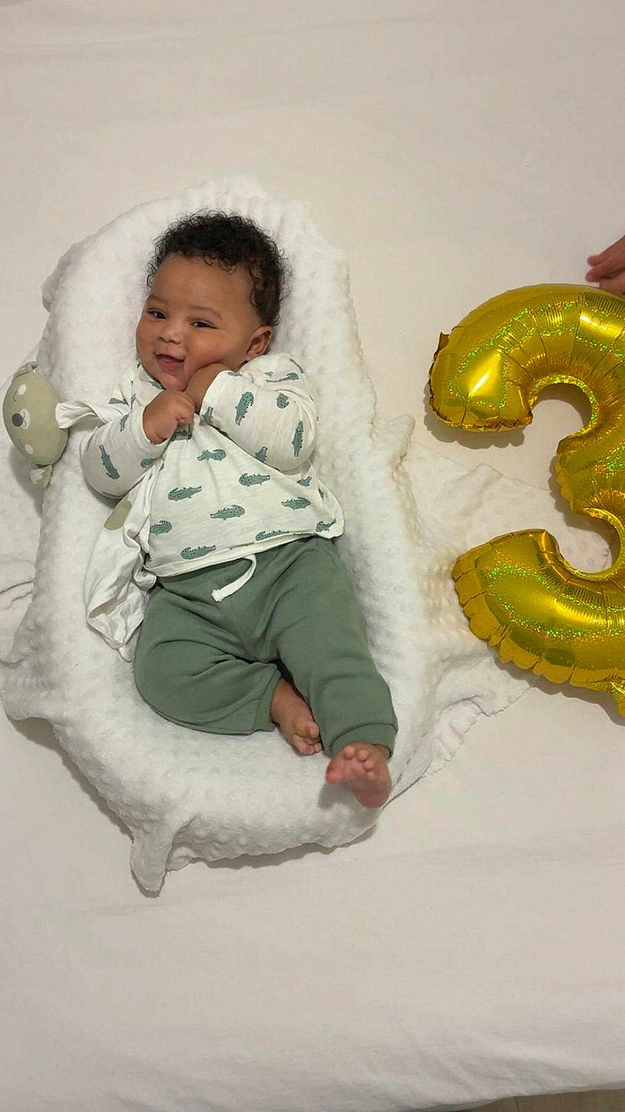 Ayden a rejoint le concours — aidez-le/la à gagner de superbes lots ! baby, infant, cushion, blanket, green_pants, crocodile_pattern, smiling, face, barefoot, balloon, number_three, celebration, hand, curly_hair, white_background, soft_texture, lying_down, cute, happy, indoors
