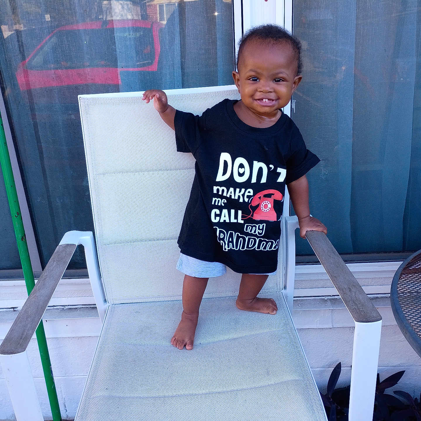 K'La is registered to the contest to win money with this photo: barefoot, black_tshirt, chair, child, clothing, daylight, happy, home, mesh_screen, outdoor, patio, person, playful, red_vehicle, shorts, smiling, standing, toddler, window, young_child