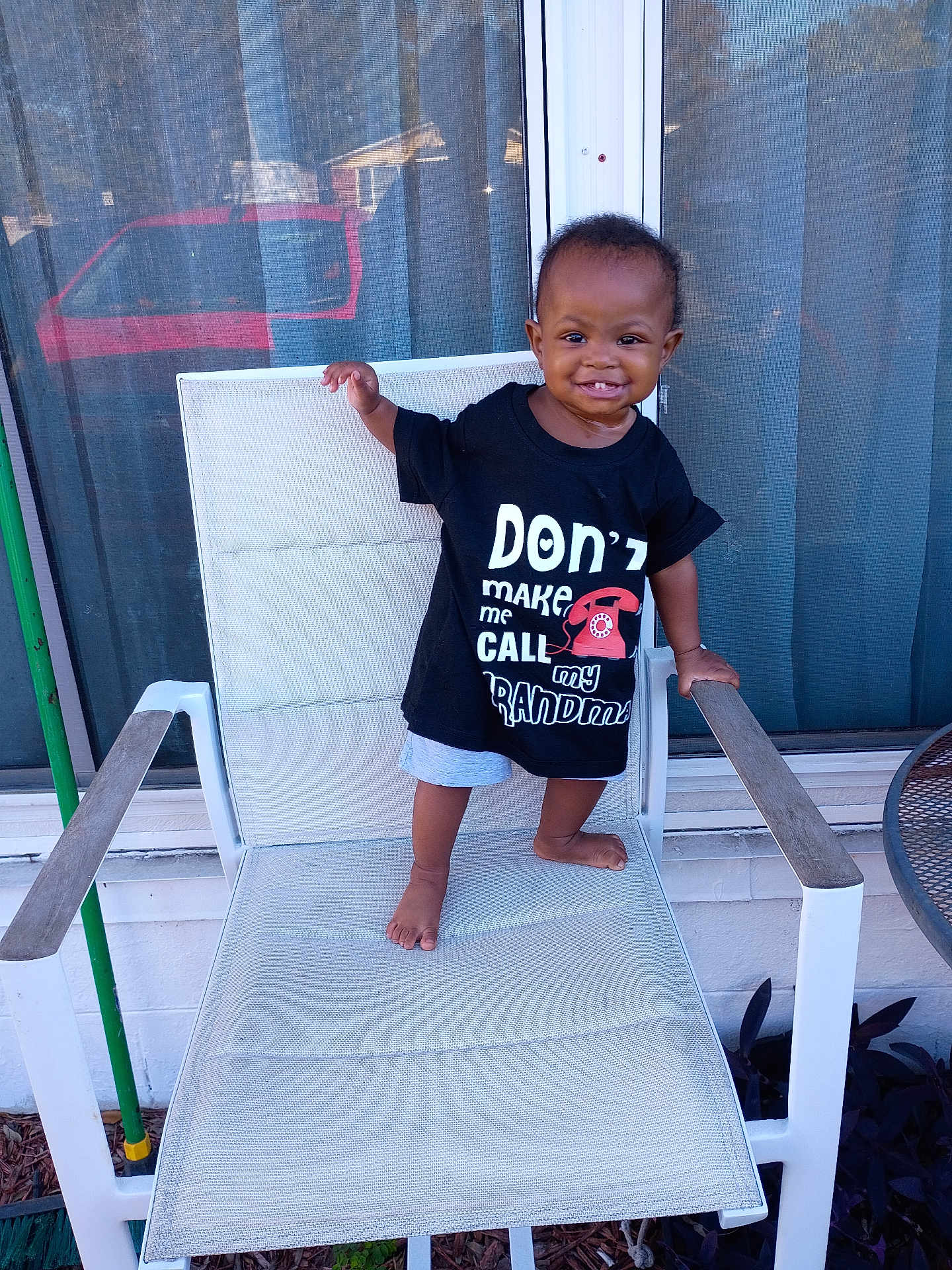 K'La is registered to the contest to win money with this photo: toddler, child, smiling, black_tshirt, barefoot, chair, patio, outdoor, window, mesh_screen, red_vehicle, happy, person, clothing, shorts, young_child, standing, home, daylight, playful