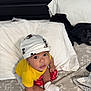 baby, child, mickey_mouse, hat, yellow_shirt, white_socks, bed, pillow, blanket, floral_pattern, indoor, cute, curious, infant, portrait, sitting, clothing, person, soft_lighting, home