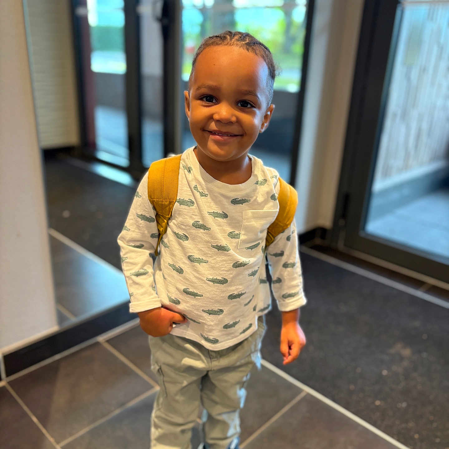 Mokeli participe au concours pour gagner de l'argent avec cette photo : child, smiling, indoor, backpack, white_shirt, crocodile_pattern, pants, sneakers, tile_floor, glass_door, natural_light, portrait, standing, young_child, happy, casual_clothing, boy, short_hair, modern_interior, daylight
