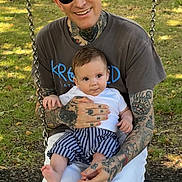 Bodhi is registered to the contest to win money with this photo: man, baby, tattoos, sunglasses, swing, outdoor, grass, trees, shirt, shorts, holding, smiling, child, nature, daylight, park, person, casual, happy, family