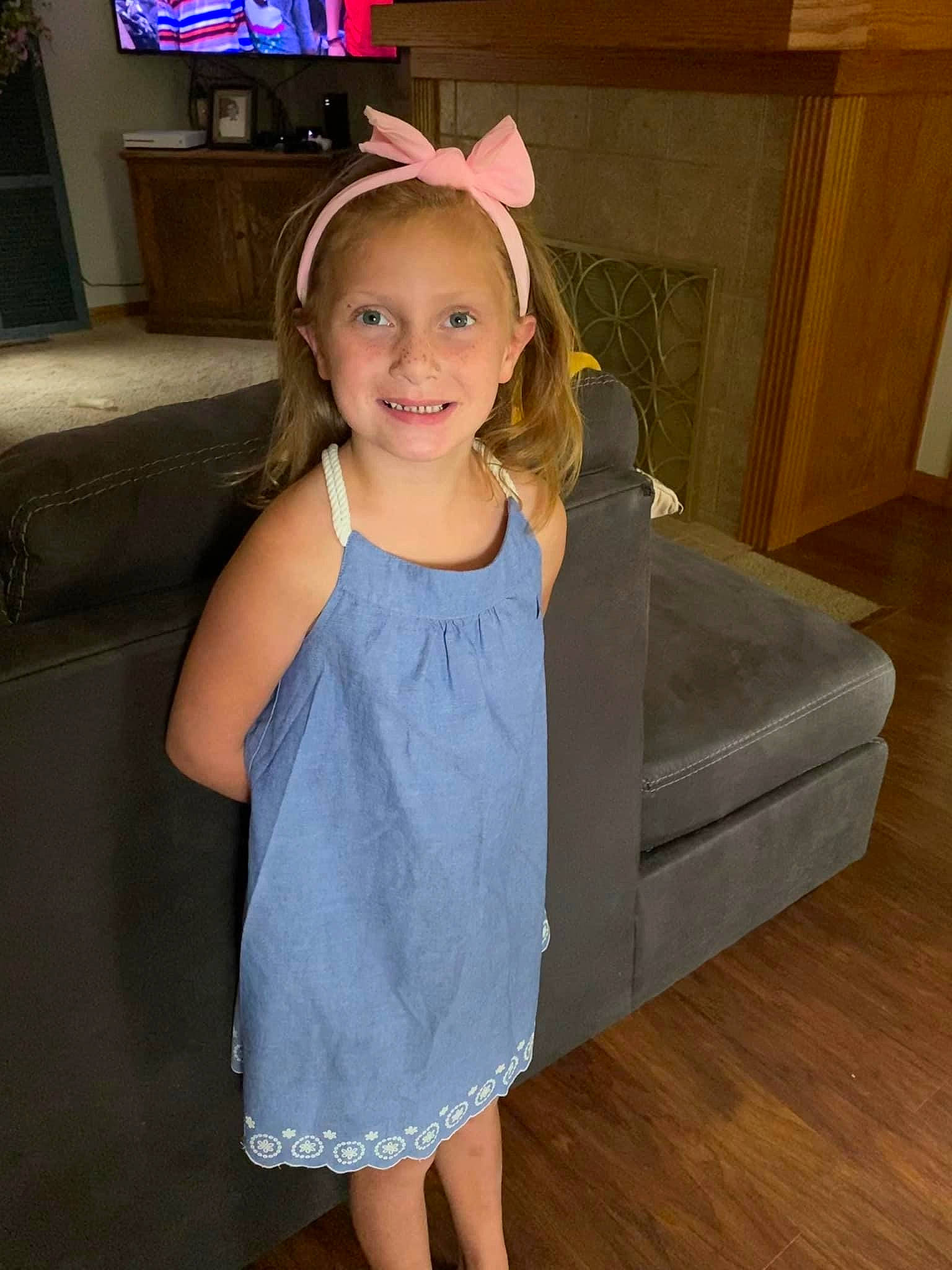 Mara is registered to the contest to win money with this photo: baby_toddler_clothing, blond, blue, couch, day_dress, dress, floor, flooring, fun, happy, hardwood, human_leg, joy, long_hair, one_piece_garment, person, sleeve, smile, television, toddler