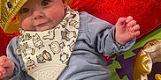 Charlie is registered to the contest to win money with this photo: baby, crown, bib, blue_clothing, toy, red_blanket, foam_mat, playroom, infant, child, cute, smiling, indoor, person, headwear, hand, floor, soft_toy, colorful, happy