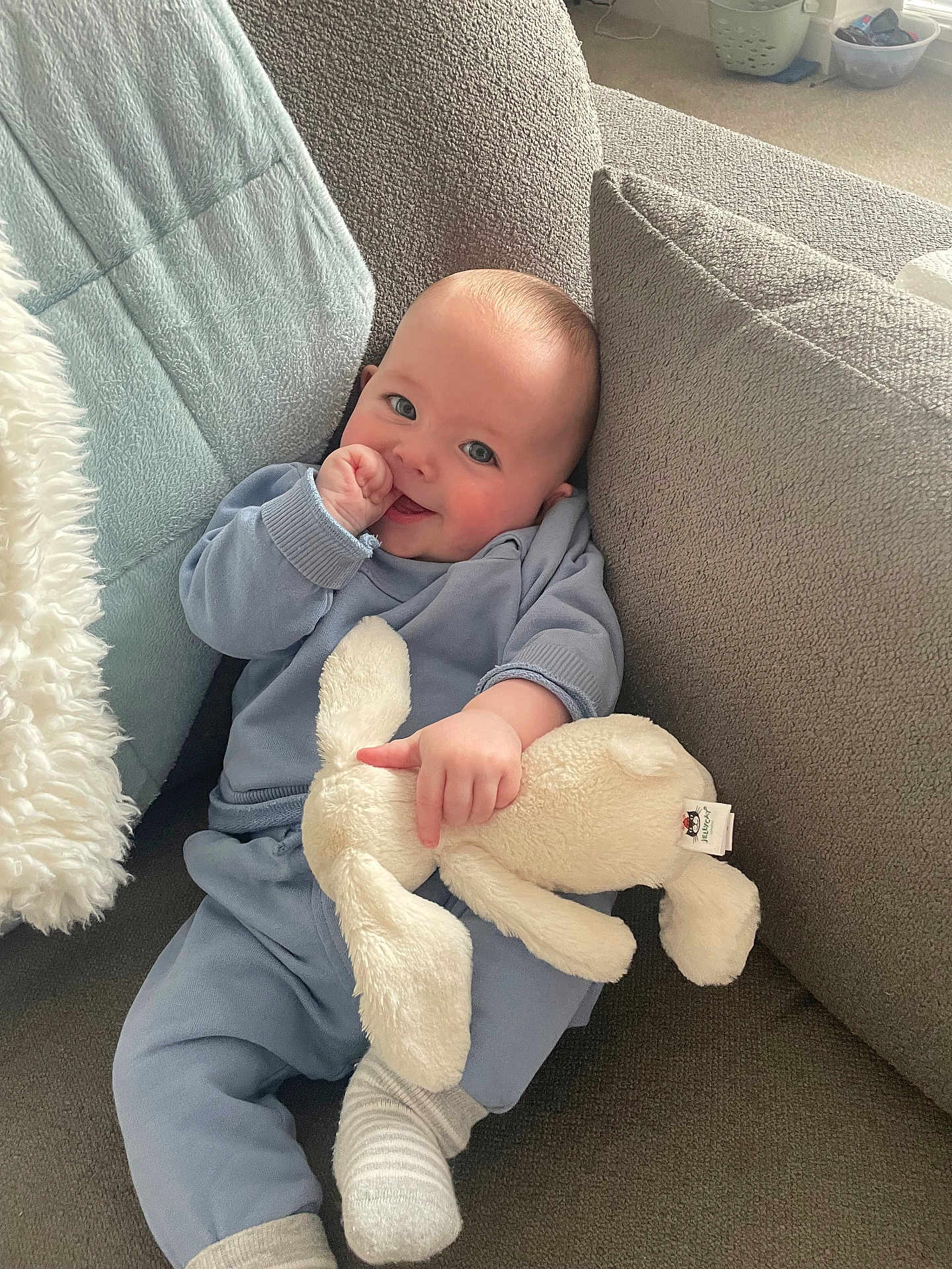 Charlie is registered to the contest to win money with this photo: baby, child, infant, blue_pajamas, stuffed_animal, bunny, couch, cushion, blanket, smiling, thumb_sucking, indoor, cozy, soft_toy, carpet, home, happy, cute, relaxed, person
