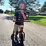 Porter is registered to the contest to win money with this photo: child, helmet, balance_bike, street, sunlight, outdoor, tree, grass, shorts, tshirt, sneakers, shadow, residential, sidewalk, summer, daytime, person, vehicle, quiet, thinking