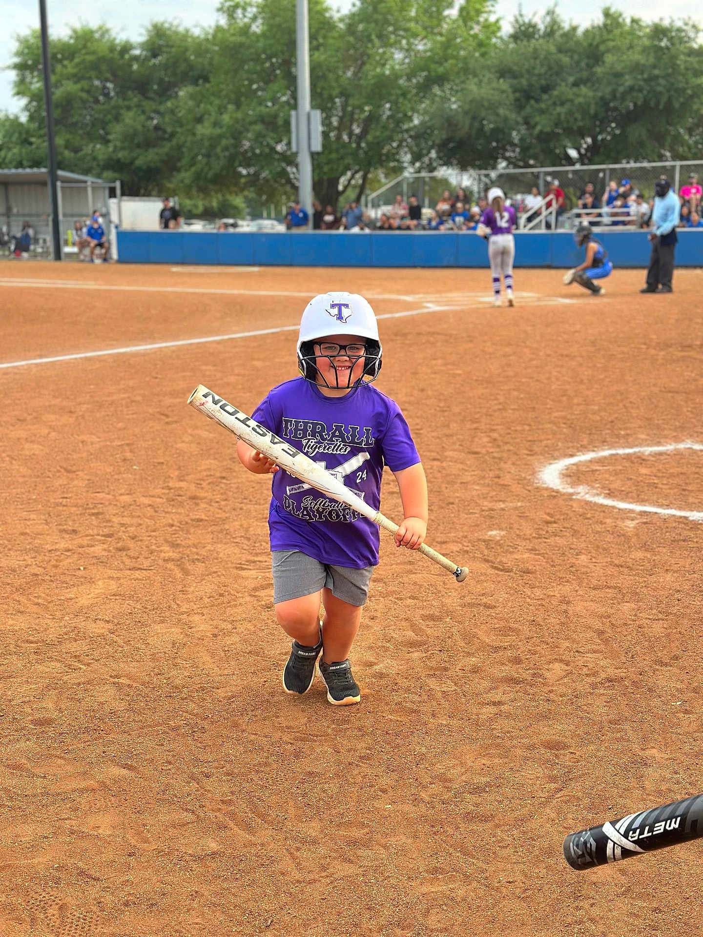 Levi is registered to the contest to win money with this photo: child, softball, baseball_bat, helmet, sports, field, running, purple_shirt, glasses, dirt, outdoor, crowd, spectators, fence, umpire, catcher, player, shorts, shoes, daytime