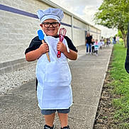 Levi joined the competition — help win amazing prizes! child, chef_hat, apron, glasses, smiling, holding_utensils, spatula, whisk, sidewalk, outdoor, concrete, building_wall, tree, grass, people, line, casual_clothing, black_shoes, shorts, happy