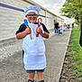 child, chef_hat, apron, glasses, smiling, holding_utensils, spatula, whisk, sidewalk, outdoor, concrete, building_wall, tree, grass, people, line, casual_clothing, black_shoes, shorts, happy