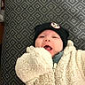 apparel, baby, child, clothing, cozy, cute, expression, face, hat, headwear, indoor, infant, jacket, lying_down, patterned_background, person, playful, smiling, warm, young_child