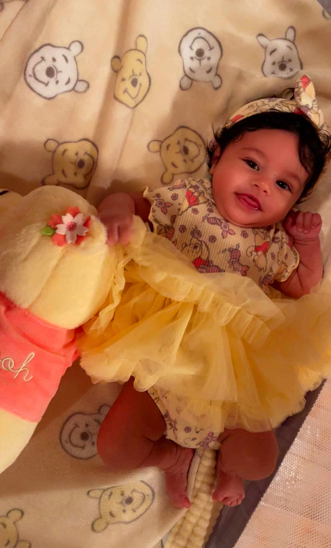 River is registered to the contest to win money with this photo: baby, infant, smiling, yellow_dress, headband, plush_toy, winnie_the_pooh, blanket, cute, lying_down, child, indoors, happy, soft_toy, patterned_clothing, feet, hand, portrait, adorable, cozy