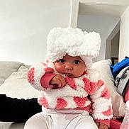 Mayannah participe au concours pour gagner de l'argent avec cette photo : baby, child, fluffy_hat, pink_and_white, sweater, couch, indoor, bottle, curious_expression, seated, cozy, living_room, white_pants, soft_texture, person, cute, infant, portrait, hat_with_bow, casual_clothing