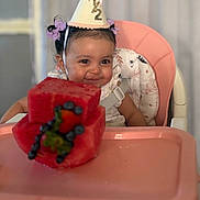 Alaiyah is registered to the contest to win money with this photo: baby, child, high_chair, party_hat, watermelon, cake, fruit, blueberries, strawberry, smile, celebration, birthday, cute, pink, food, face, person, happy, indoors, toddler