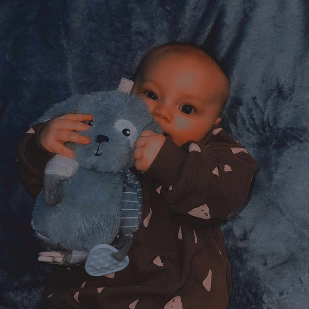 Wylder is registered to the contest to win money with this photo: baby, blanket, blue, child, clothing, cute, face, hands, holding, indoor, infant, jeans, lying_down, person, plush, soft_texture, stuffed_animal, sweater, toy, young