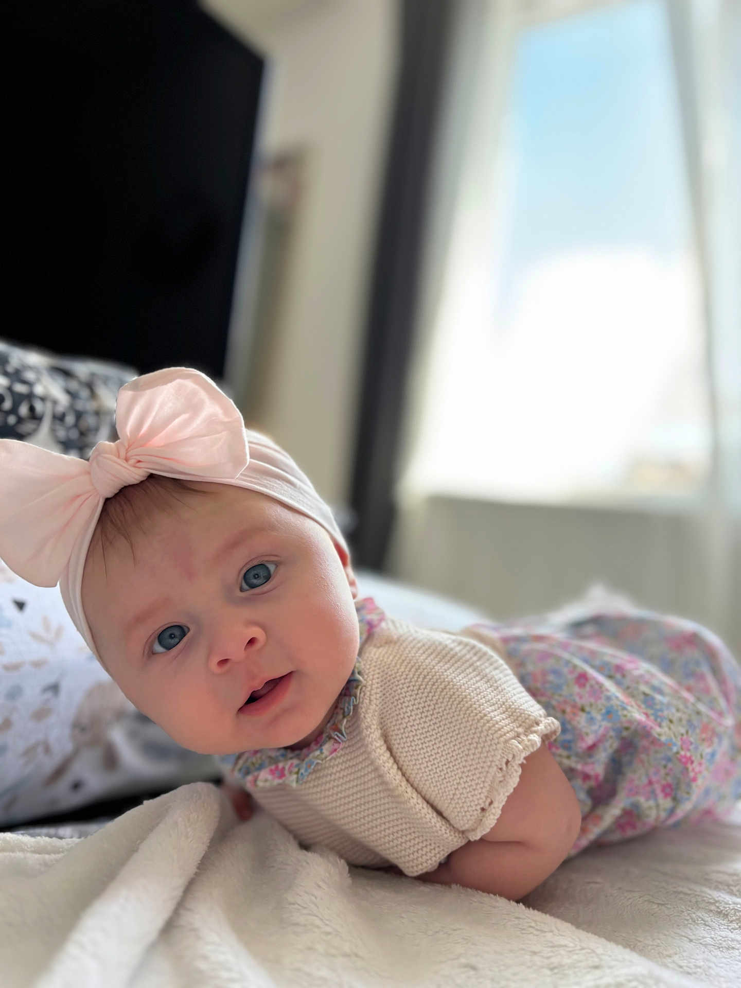 Heïly participe au concours pour gagner de l'argent avec cette photo : baby, blanket, blue_eyes, clothing, curious, cute, expression, face, headband, indoors, infant, knitwear, lying_down, natural_light, person, pink_bow, portrait, soft_texture, toddler, window