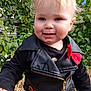 black_clothing, blonde_hair, child, curly_hair, cute, fall, greenery, happy, leather_jacket, leaves, nature, outdoor, person, portrait, red_bow, sequins, sitting, smiling, toddler, zipper
