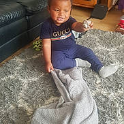 Tyler is registered to the contest to win money with this photo: blanket, carpet, child, clothing, curly_hair, expression, floor, furniture, indoor, living_room, pacifier, person, play, plush_toy, rug, sitting, smiling, sofa, toddler, toy