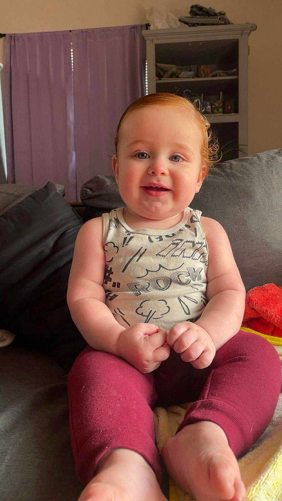 Marvin is registered to the contest to win money with this photo: baby, child, infant, couch, clothing, sleeveless_shirt, pants, red_hair, blue_eyes, smiling, indoor, furniture, curtains, cabinet, blanket, home, person, portrait, sitting, cute