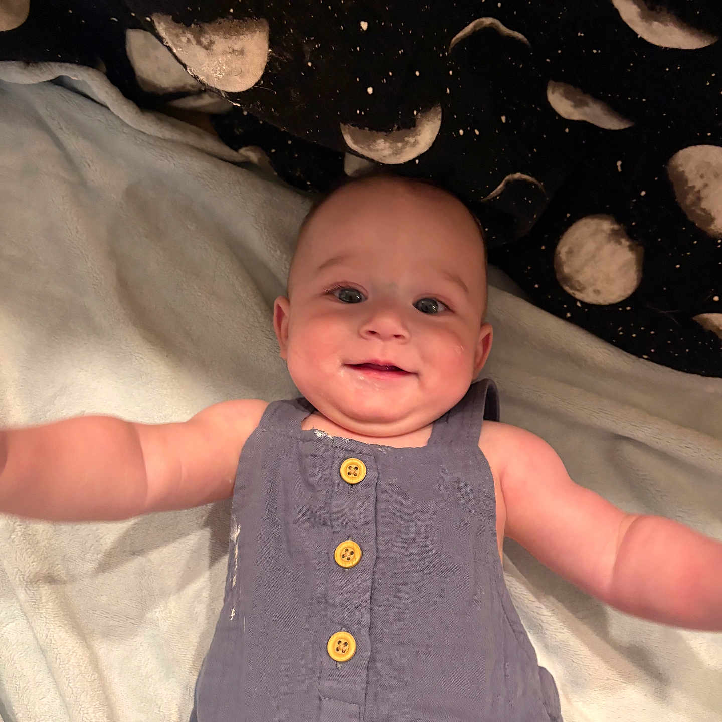 Braxton is registered to the contest to win money with this photo: arms_outstretched, baby, blanket, blue_clothing, buttoned_clothing, child, comfort, cute, eyes, face, happy, head, indoors, infant, lying_down, moon_pattern, romper, skin, smiling, soft_texture