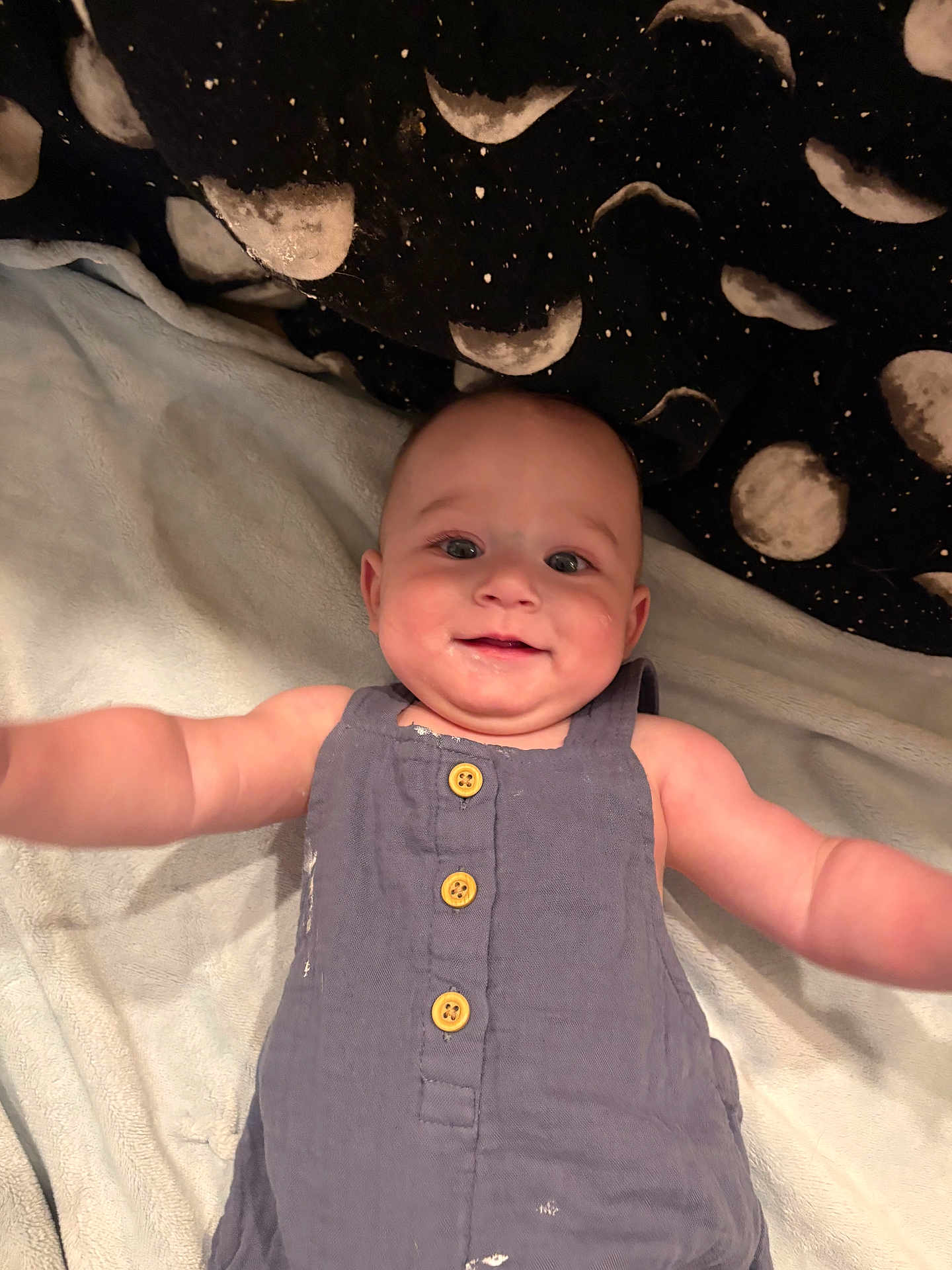 Braxton is registered to the contest to win money with this photo: baby, infant, smiling, romper, blue_clothing, blanket, moon_pattern, lying_down, arms_outstretched, happy, cute, child, face, head, eyes, skin, buttoned_clothing, indoors, soft_texture, comfort