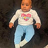 baby, child, sitting, white_shirt, colorful_text, blue_pants, socks, black_rug, floor, indoor, cute, portrait, calm, curious, infant, young_child, clothing, person, human, soft_texture