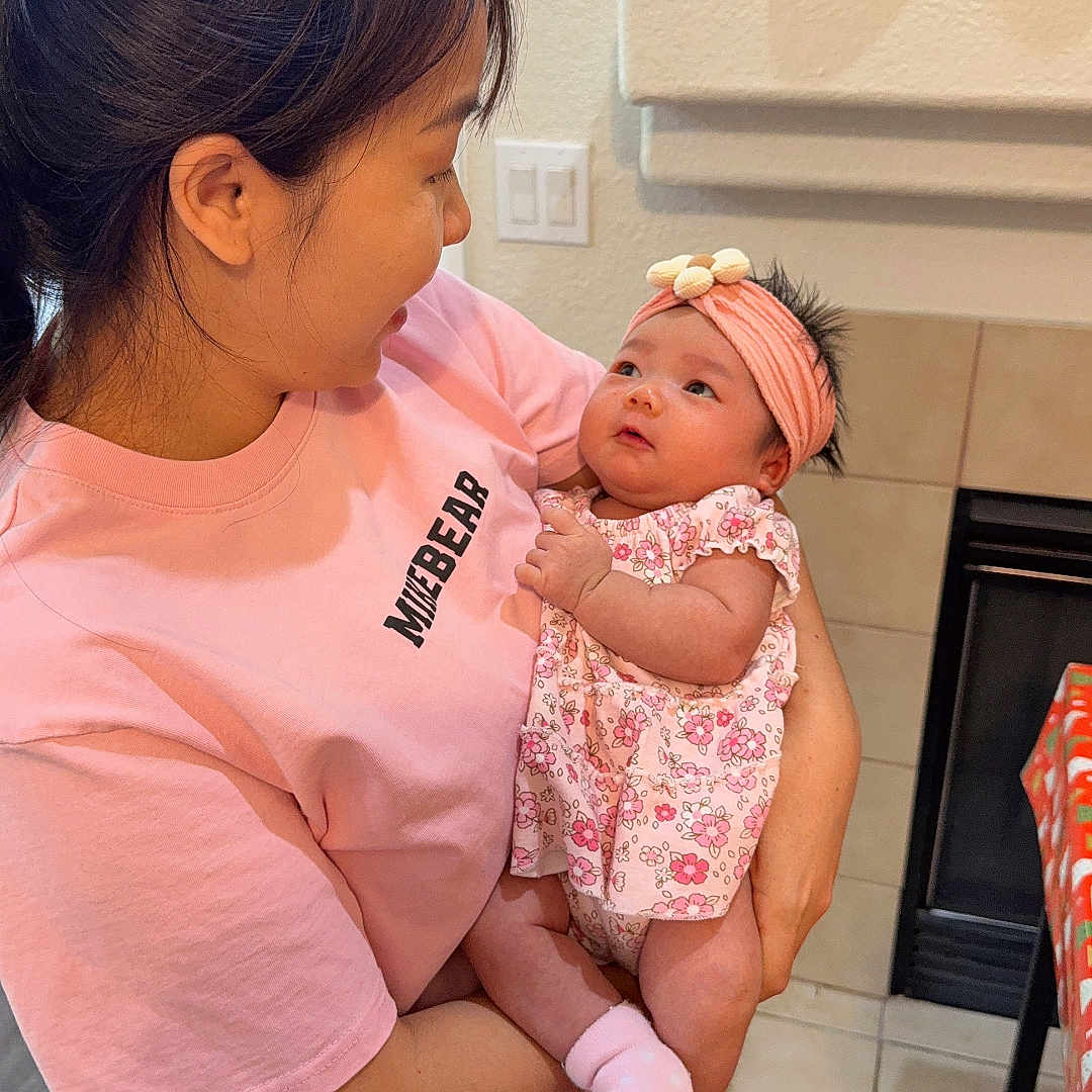 Selena is registered to the contest to win money with this photo: affection, baby, family, fireplace, floor, floral_dress, fruit, grapes, headband, holding, indoor, infant, kitchen, light_switch, pink_shirt, socks, table, tile, woman, wood_floor