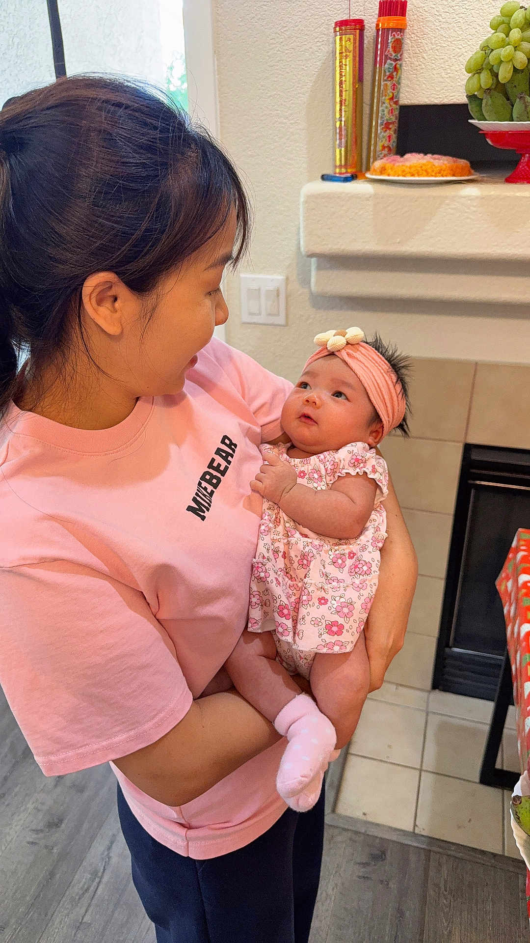 Selena is registered to the contest to win money with this photo: woman, baby, infant, holding, pink_shirt, headband, floral_dress, socks, indoor, kitchen, table, fruit, grapes, fireplace, floor, tile, wood_floor, light_switch, affection, family