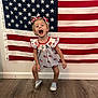 Madelyn is registered to the contest to win money with this photo: child, girl, american_flag, dress, hair_bows, smiling, shoes, wooden_floor, indoor, celebration, patriotic, happy, person, wall, necklace, white_wall, patterned_dress, standing, joyful, cute