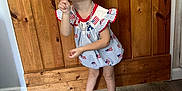 Madelyn is registered to the contest to win money with this photo: child, girl, peace_sign, pucker_lips, patriotic, dress, headband, curly_hair, standing, wooden_background, floor, shoes, casual, indoor, necklace, playful, cute, pose, person, happy