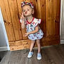 child, girl, peace_sign, pucker_lips, patriotic, dress, headband, curly_hair, standing, wooden_background, floor, shoes, casual, indoor, necklace, playful, cute, pose, person, happy