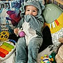 baby, child, dinosaur_onesie, plush_toy, colorful_toys, crib, pillow, cute, indoor, soft_toys, playtime, smiling, hand_in_mouth, infant, stuffed_animal, floor_mat, cozy, comfort, young_child, playroom