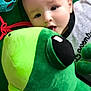 Arès participe au concours pour gagner de l'argent avec cette photo : baby, plush_toy, green, dinosaur, infant, play, toy, child, cute, lying_down, hand, face, soft_toy, colorful, indoors, happy, young, childhood, smiling, clothing