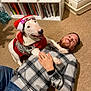 dog, man, carpet, santa_hat, sweater, indoor, bookshelf, books, smiling, happy, casual_clothing, beard, plaid_shirt, watch, paw, embrace, home, floor, festive, relaxing