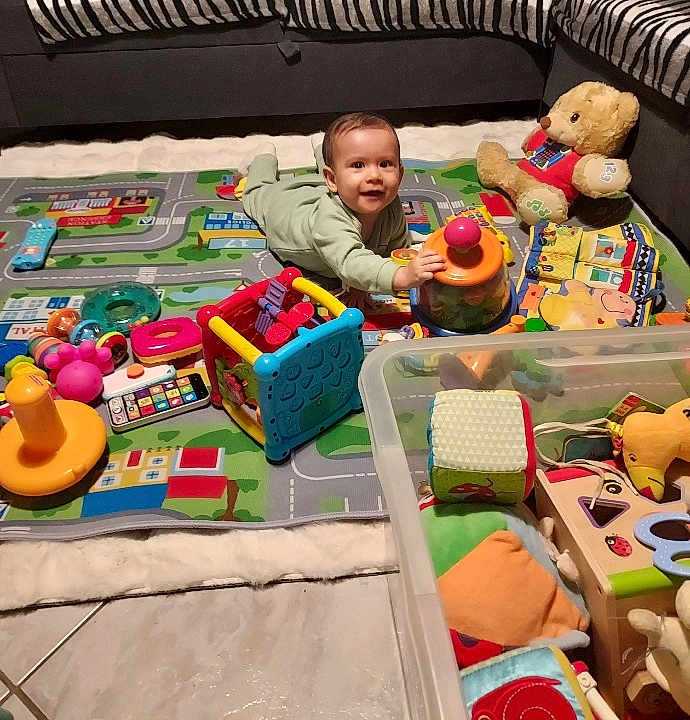 Julio participe au concours pour gagner de l'argent avec cette photo : baby, toddler, toy, play_mat, living_room, couch, teddy_bear, child, floor, colorful, plastic_toys, soft_toys, playtime, smiling, indoors, cushion, puzzle_toy, clothing, person, happy
