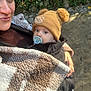 baby, pacifier, hat, pom_poms, blanket, adult, smile, outdoor, nature, greenery, person, portrait, warm_clothing, stone_wall, hand, nail_polish, face, cute, winter_wear, cozy