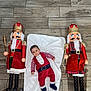 baby, child, nutcracker, toy, holiday, christmas, red_outfit, blanket, wooden_floor, decor, festive, face, person, indoor, cute, lying_down, blue_eyes, white_beard, mustache, smile