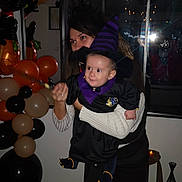 Julio a rejoint le concours — aidez-le/la à gagner de superbes lots ! baby, balloons, black_balloons, celebration, child, costume, decorations, festive, floor, halloween_costume, holding, indoor, orange_balloons, party, person, reflection, smiling, window, witch_hat, woman