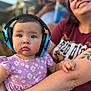 Mia is registered to the contest to win money with this photo: baby, headphones, child, woman, tattoo, purple_dress, floral_pattern, outdoor, portrait, holding, smiling, face, person, people, blurred_background, arm, casual_clothing, happy, family, daylight