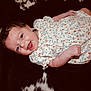 Oakley is registered to the contest to win money with this photo: baby, smiling, infant, floral_dress, happy, lying_down, skin, arm, hand, face, person, cute, child, indoors, closeup, black_and_white_background, soft_texture, cheerful, young, portrait