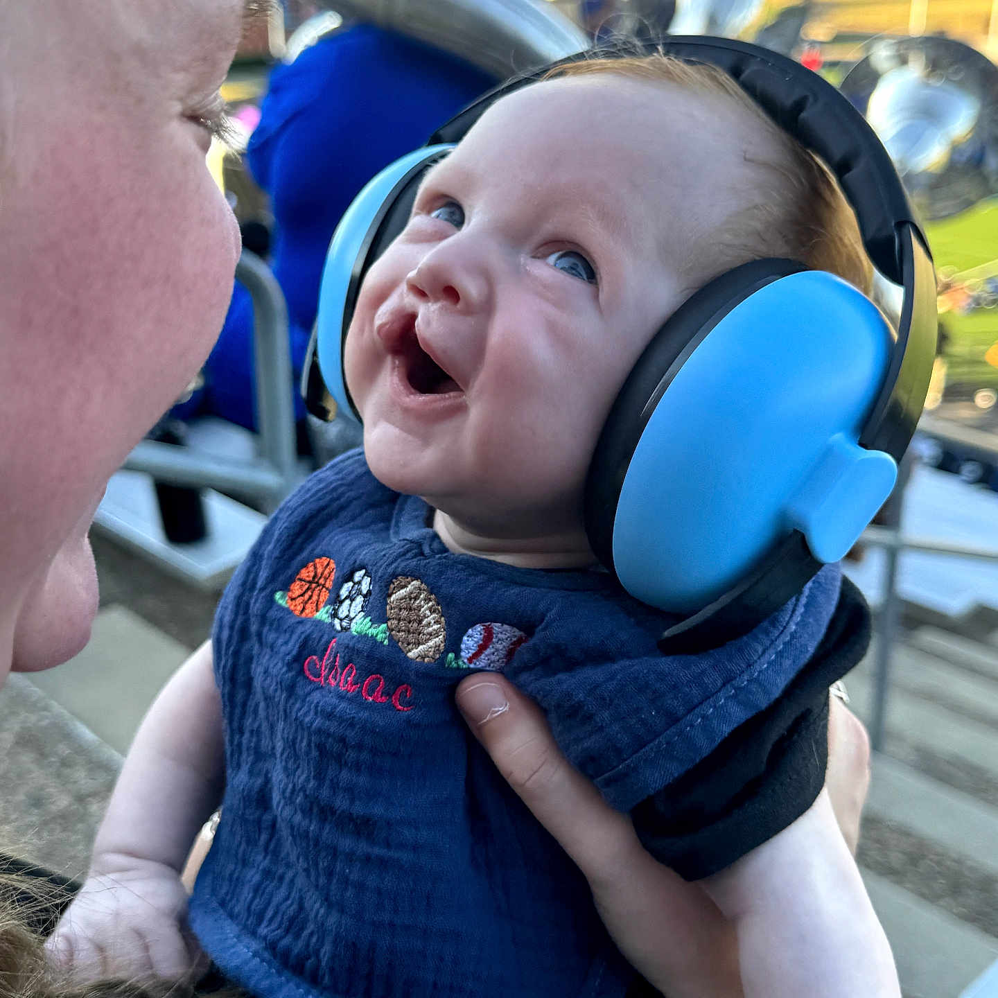 Isaac is registered to the contest to win money with this photo: baby, infant, child, smiling, happy, hearing_protection, earmuffs, blue, portrait, close_up, adult, parent, outdoor, daylight, amusement_park, seated, held, clothing, face, family