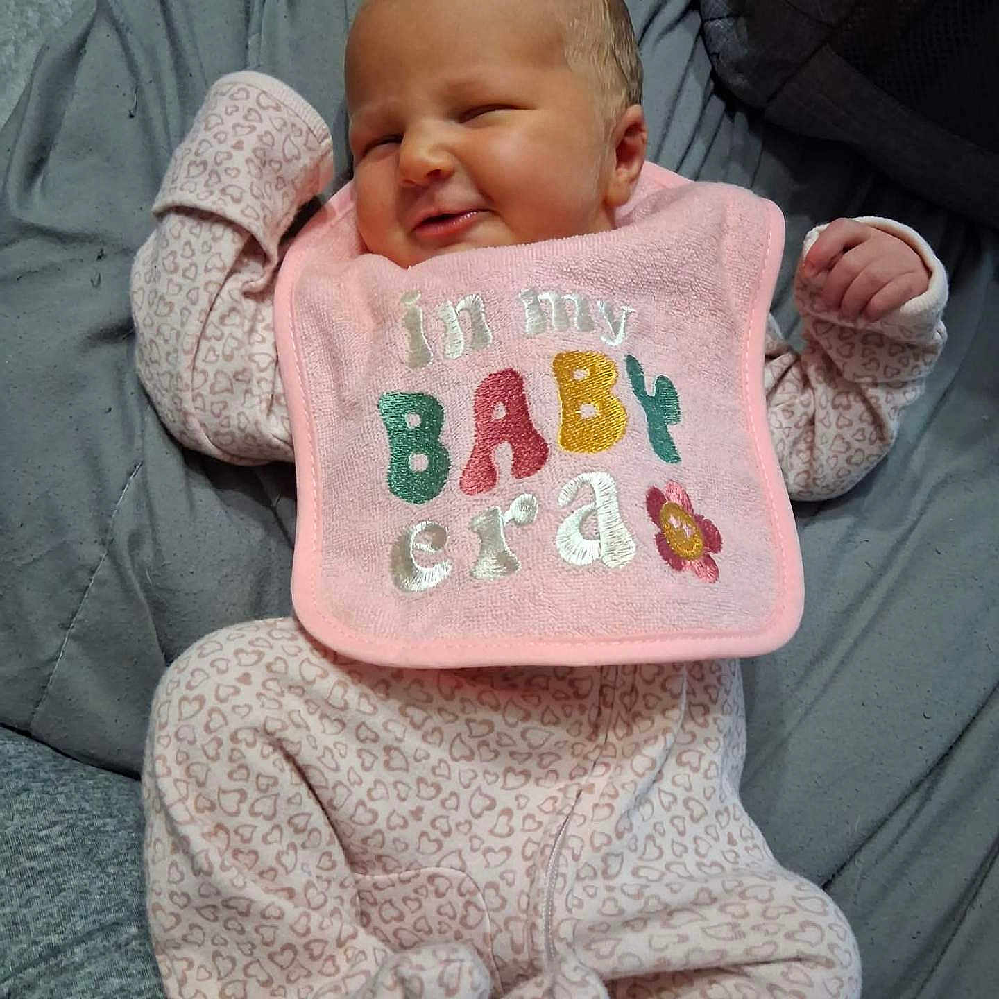 Daveena is registered to the contest to win money with this photo: baby, infant, onesie, bib, pink, smile, face, cheeky, heart_pattern, clothing, blanket, sleepwear, newborn, cute, person, lying_down, soft_texture, indoor, child, happy