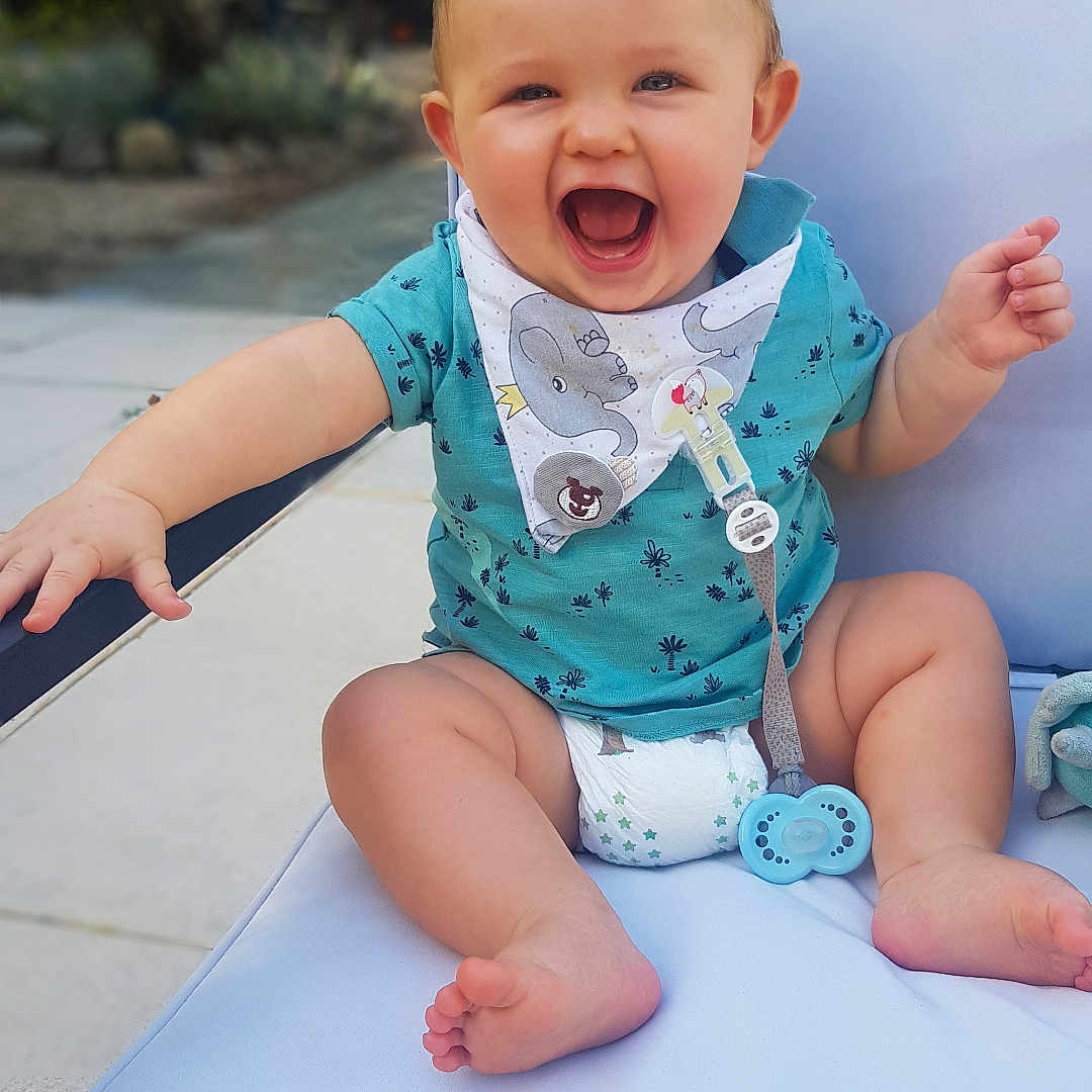 Léo participe au concours pour gagner de l'argent avec cette photo : baby, barefoot, bib, blue_clothing, child, cushion, cute, diaper, garden, happy, infant, laughing, outdoor, pacifier, person, playful, seat, smiling, summer, sunlight