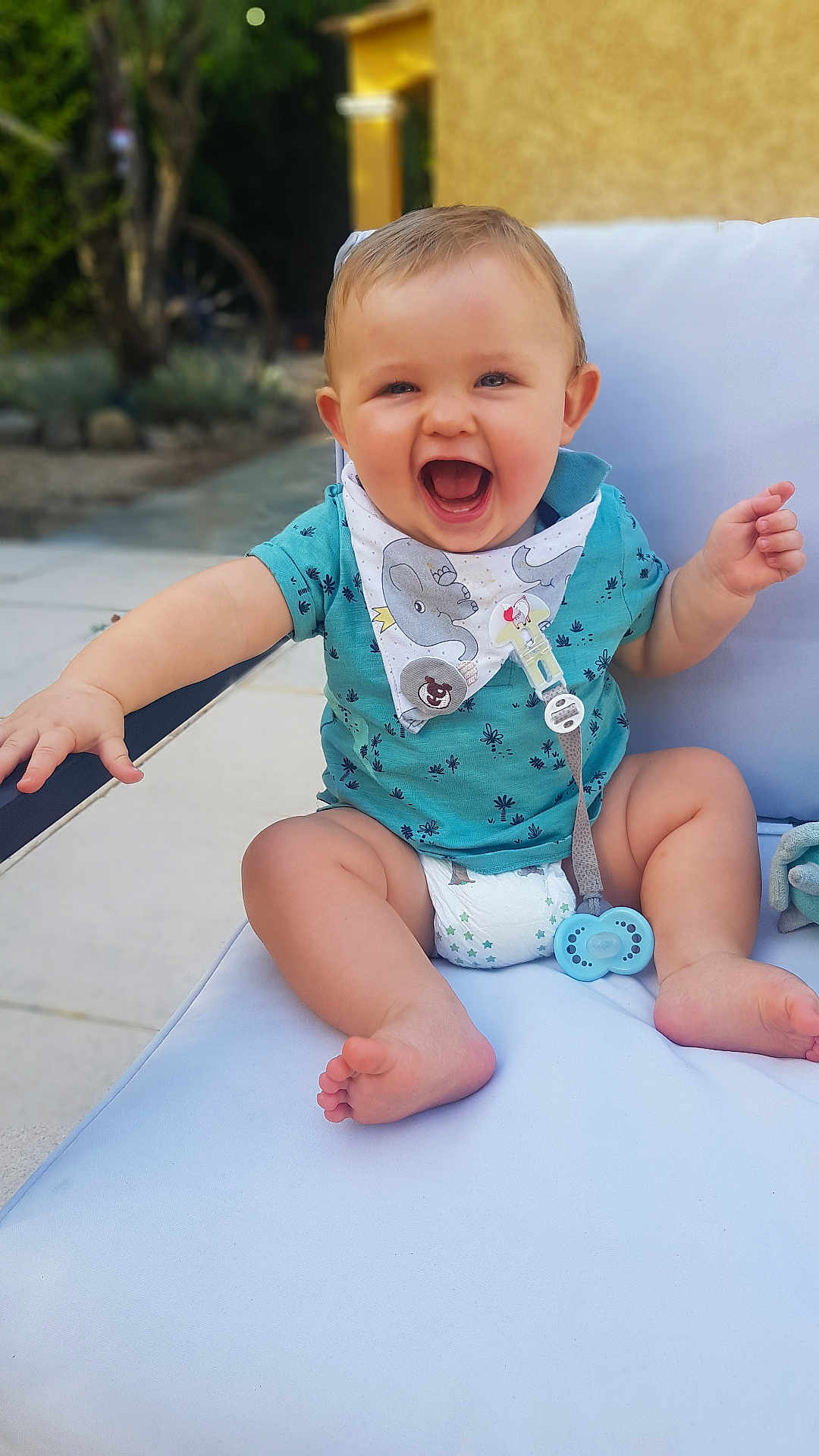 Léo participe au concours pour gagner de l'argent avec cette photo : baby, smiling, laughing, child, outdoor, seat, cushion, bib, diaper, pacifier, happy, cute, person, infant, blue_clothing, summer, sunlight, barefoot, playful, garden