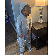 Jaquale is registered to the contest to win money with this photo: child, hooded_clothing, furry_outfit, indoor, lamp, side_table, wood_flooring, curious_expression, standing, casual_shoes, modern_furniture, neutral_colors, lighting, young_person, portrait, floor, home_interior, cozy, smile, person