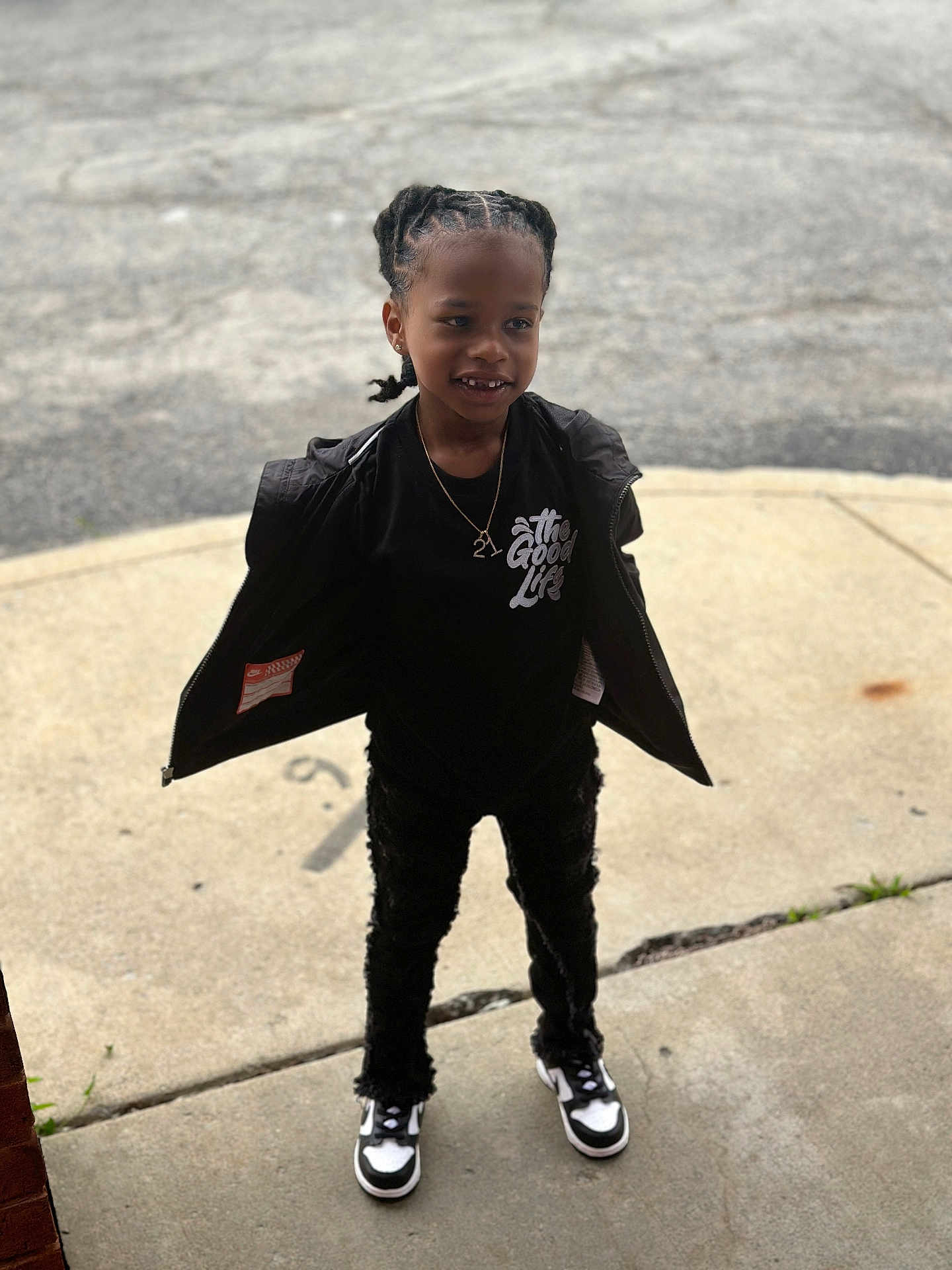 Jaquale is registered to the contest to win money with this photo: child, outdoor, sidewalk, black_jacket, black_shirt, white_text, braided_hair, smile, black_pants, sneakers, concrete, standing, person, young_child, casual_clothing, fashion, street, portrait, daylight, happy