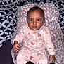 baby, infant, child, onesie, blanket, pillow, bed, patterned_bedspread, pink_clothing, heart_pattern, big_eyes, staring, sitting, lying_down, chubby_cheeks, hand, foot, portrait, indoor, cozy